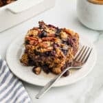 A slice of baked oatmeal with blueberries and pecans sits on a white plate with a fork, next to a striped napkin and a cup of coffee on a marble surface—perfect for enjoying your morning baked oatmeal ritual.
