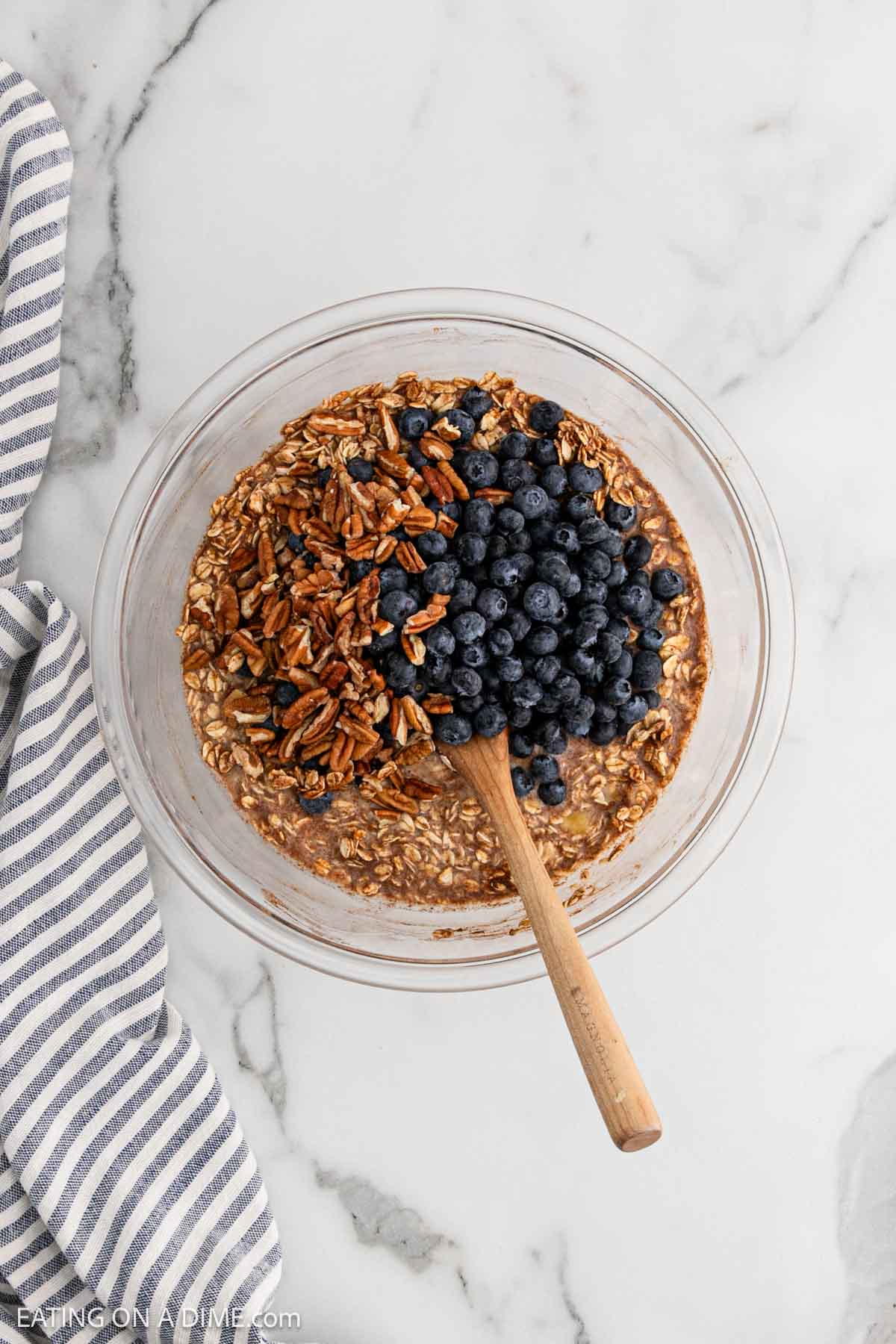 Adding in blueberries and chopped pecans into the oat mixture in a bowl with a wooden spoon