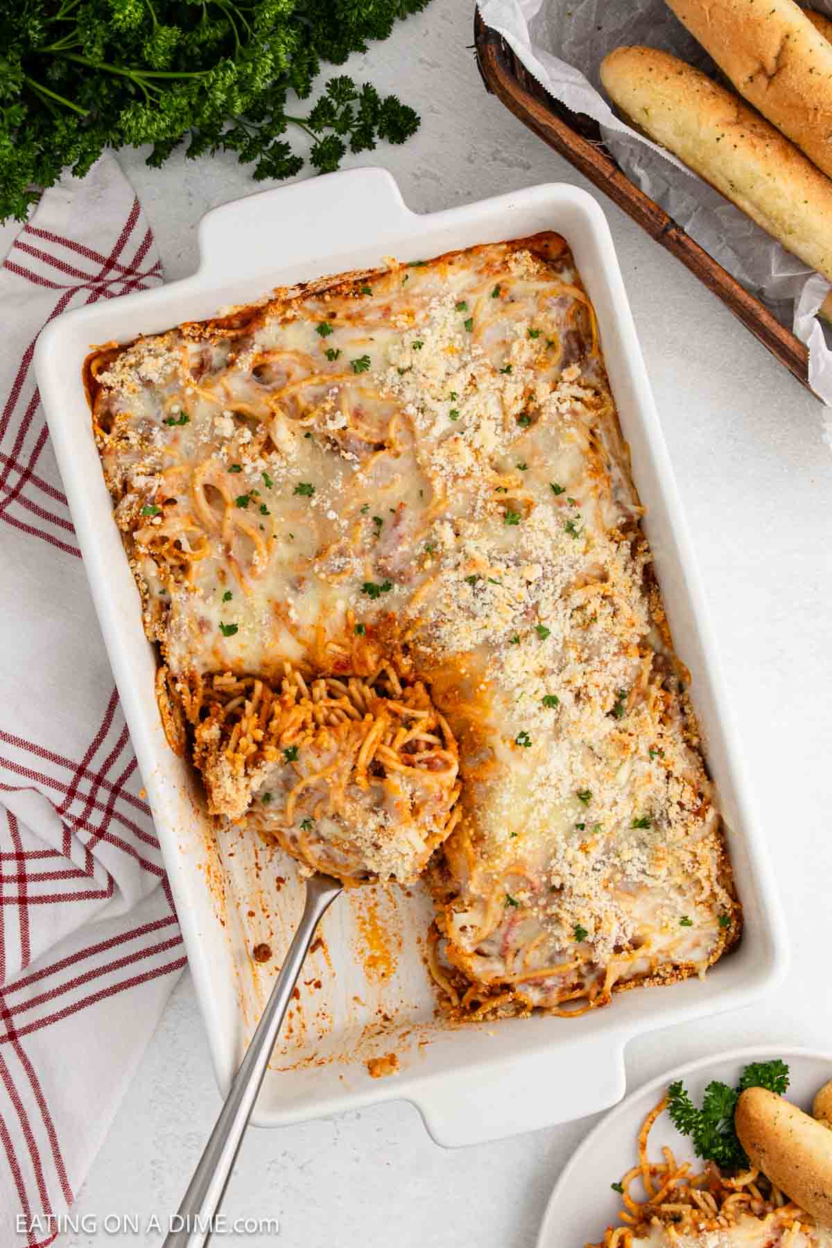 Baked Spaghetti in a casserole dish topped with melted cheese with a serving on a spatula with a side of breadsticks in a basket