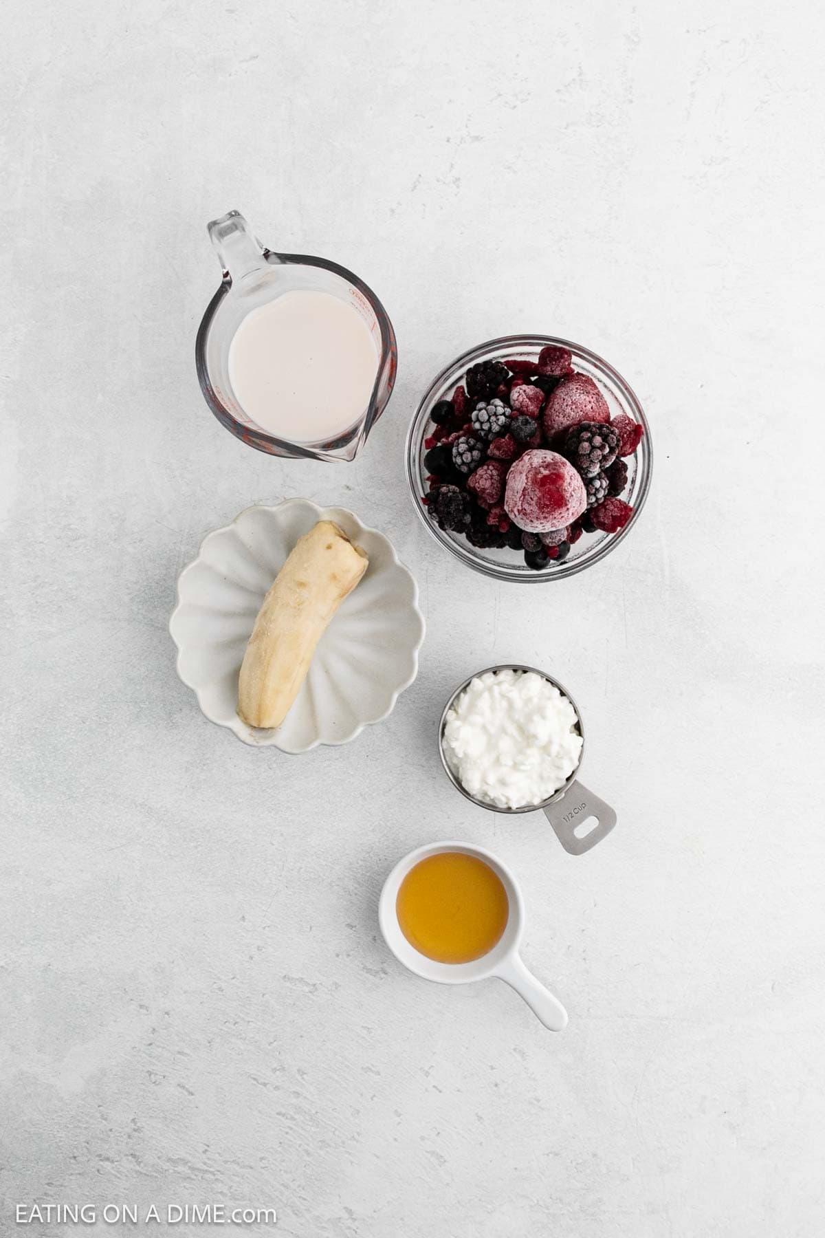 A flat lay of Cottage Cheese Smoothie ingredients: a glass measuring cup of milk, a bowl of frozen mixed berries, half a banana on a plate, cottage cheese in a measuring cup, and honey on a white background.