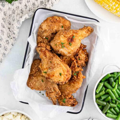 A tray of crispy Fried Chicken pieces garnished with parsley, surrounded by bowls of green beans, mashed potatoes, and corn on the cob on a white surface.
