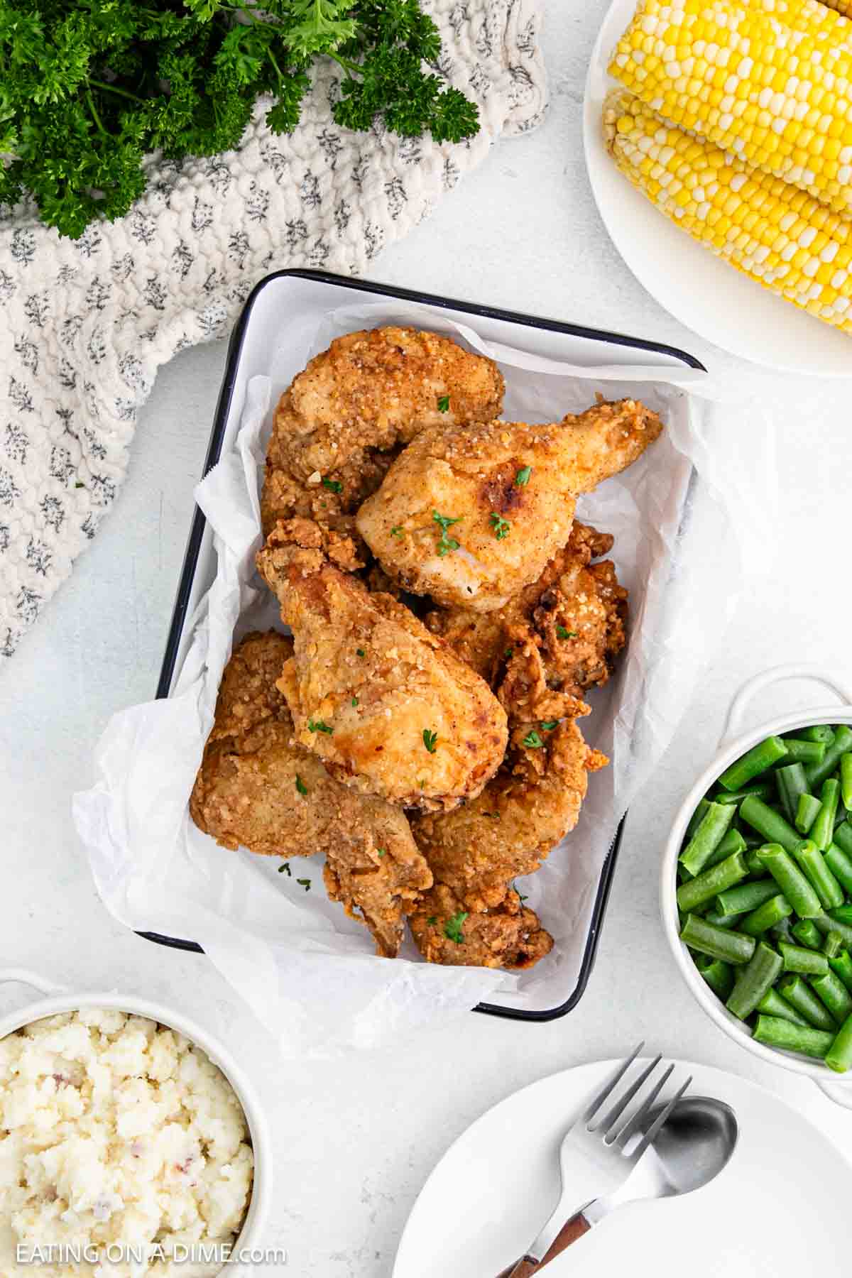 Fried Chicken placed in a parchment paper lined baking dish with a side of corn on the cob, green beans and mashed potatoes