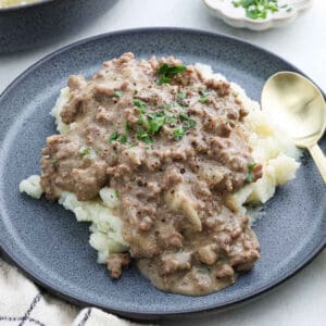 A plate of mashed potatoes topped with creamy ground beef stew and rich gravy, garnished with chopped herbs. A gold spoon is placed beside the dish on the gray plate, perfect for diving into this comforting recipe.