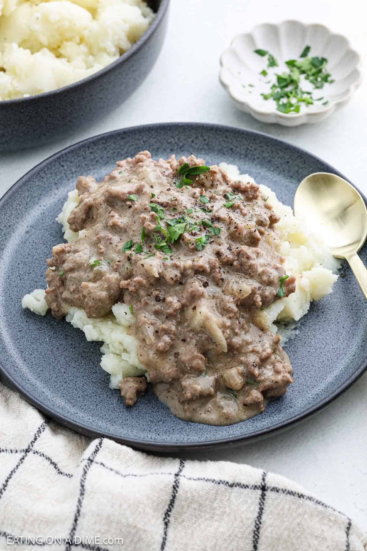 A plate of mashed potatoes topped with creamy ground beef stroganoff and rich gravy, garnished with parsley. A spoon rests beside the dish, and a small bowl of chopped herbs is nearby. A checkered cloth is partially visible in the corner.
