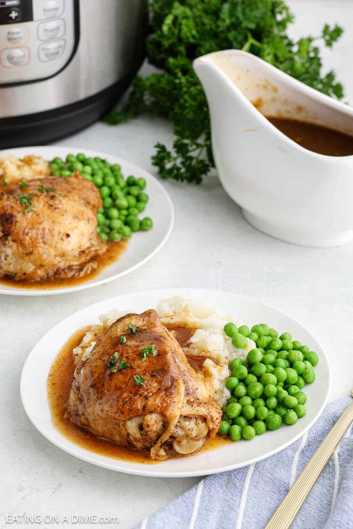 Cooked chicken thighs on a plate with gravy poured over the top on a plate with mashed potatoes and peas with a gravy bowl and a instant pot on the side