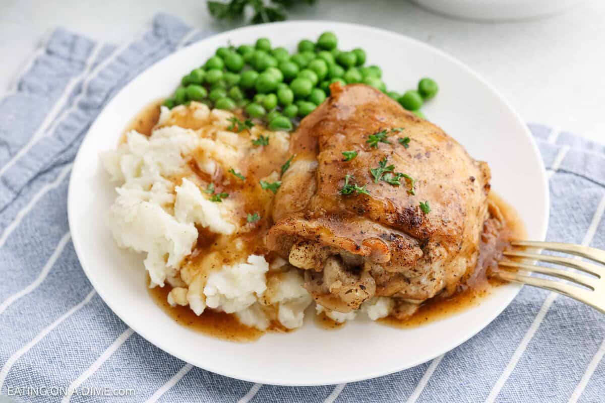 Cooked chicken thighs on a plate topped with brown gravy with a side of mashed potatoes and peas