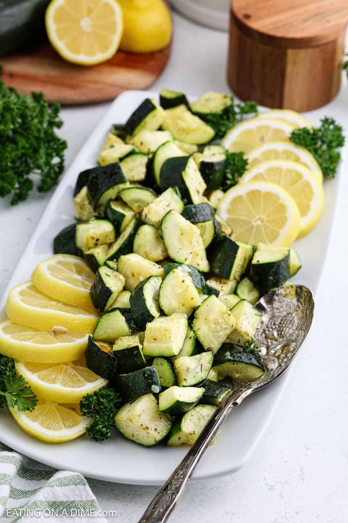 A white rectangular plate showcases roasted zucchini, diced and sprinkled with herbs. Slices of lemon and parsley elegantly garnish the dish. A silver spoon rests on the side, while more lemon and parsley can be seen in the background, making this a delightful addition to zucchini recipes.