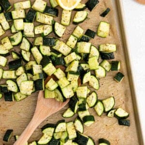 Roasted zucchini cubes, seasoned with pepper and herbs, are spread across a parchment-lined baking sheet. A wooden spoon stirs through the mix, while a lemon wedge peeks from the corner.