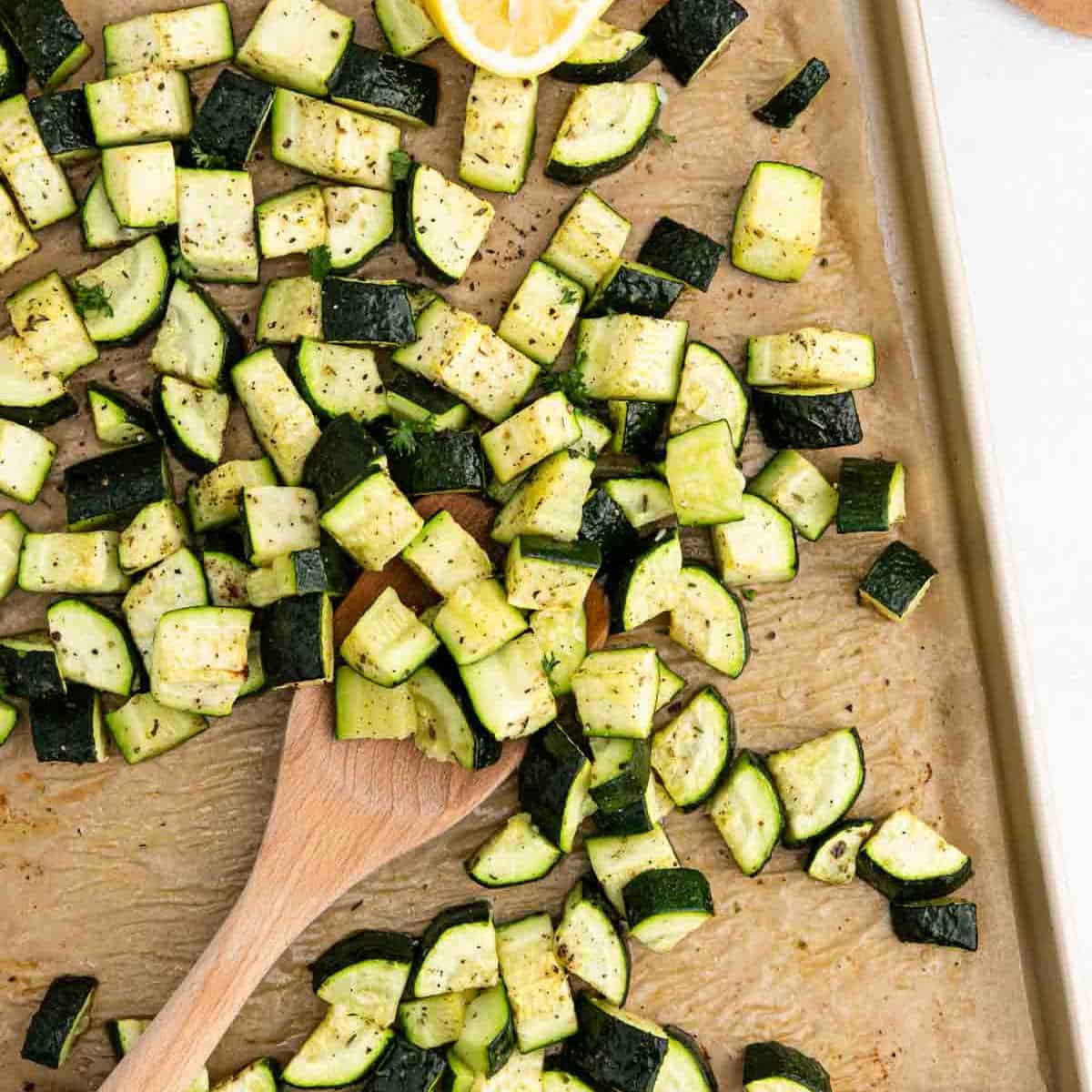 Roasted zucchini cubes, seasoned with pepper and herbs, are spread across a parchment-lined baking sheet. A wooden spoon stirs through the mix, while a lemon wedge peeks from the corner.