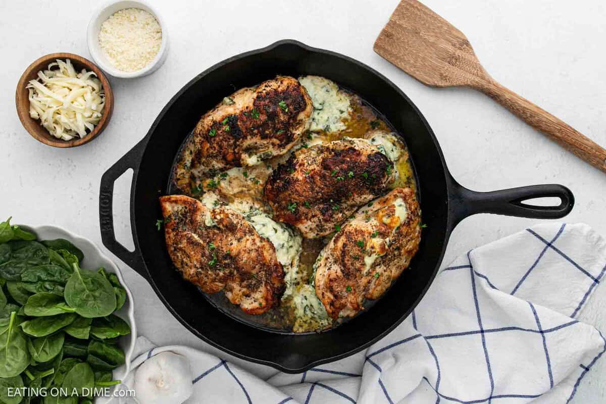 A black cast iron skillet holds four Spinach Stuffed Chicken Breasts, seasoned and filled with creamy cheese mixture, garnished with herbs. Nearby are bowls of grated cheeses, fresh spinach, and a wooden spatula on a white cloth.