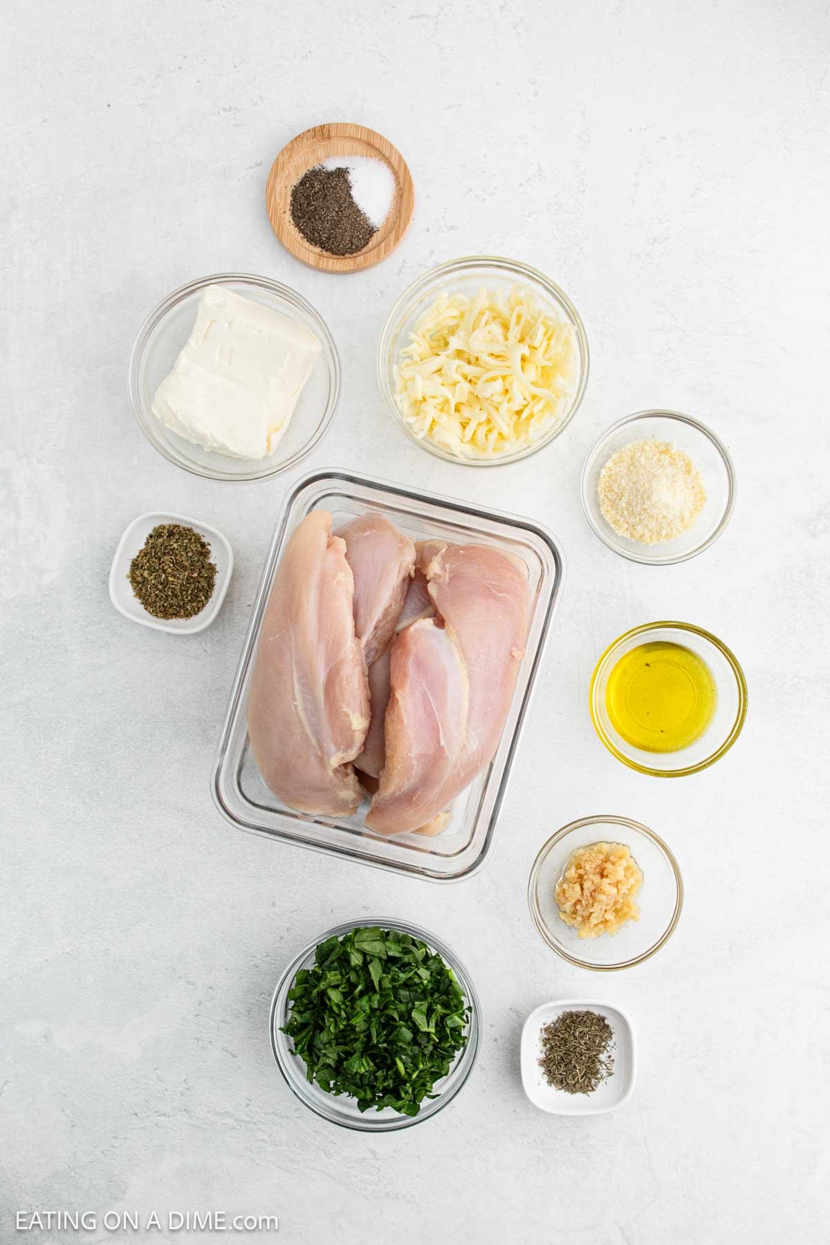 Overhead view of raw chicken breasts in a tray, ready for a delicious Spinach Stuffed Chicken Breast, surrounded by small bowls of cream cheese, shredded cheese, seasoning, breadcrumbs, olive oil, minced garlic, parsley, and black pepper.