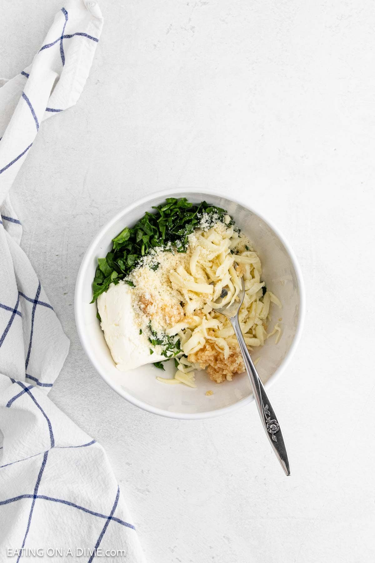 A white bowl with shredded cheese, chopped spinach, grated parmesan, cream cheese, and minced garlic sits ready for your next Spinach Stuffed Chicken recipe. A white and blue checked kitchen towel is partially visible on the white surface.