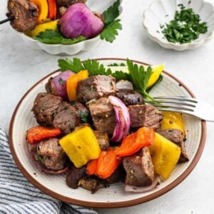 A plate of grilled steak kabobs with colorful bell peppers, red onions, and mushrooms garnished with parsley reminds you of a perfect dish from Meal Plan 50. A fork rests on the plate beside a striped napkin, while a small dish of chopped herbs sits in the background.