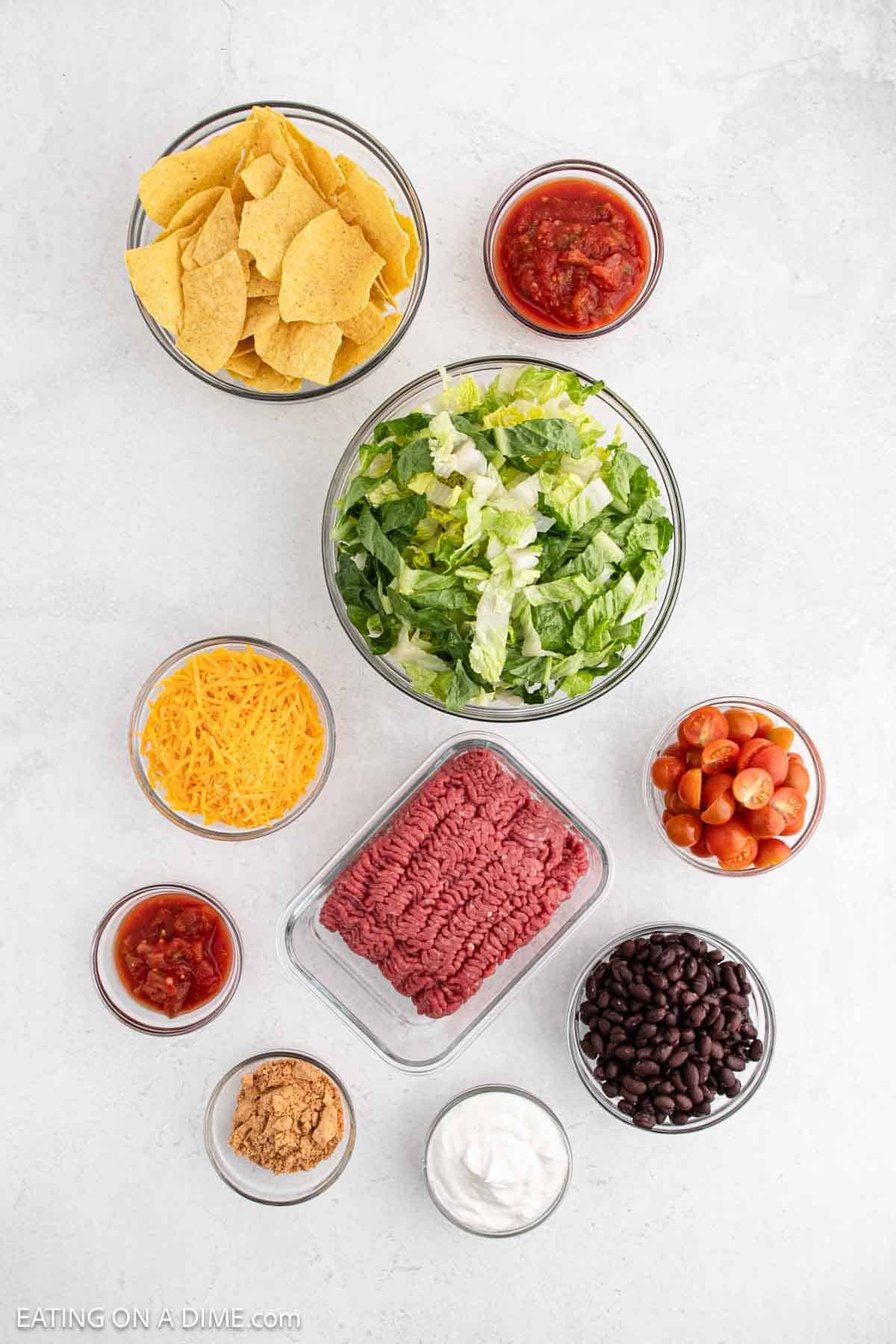 Ingredients arranged on a white background include tortilla chips, salsa, shredded cheddar cheese, ground meat, sour cream, black beans, cherry tomatoes, and a large bowl of chopped lettuce.