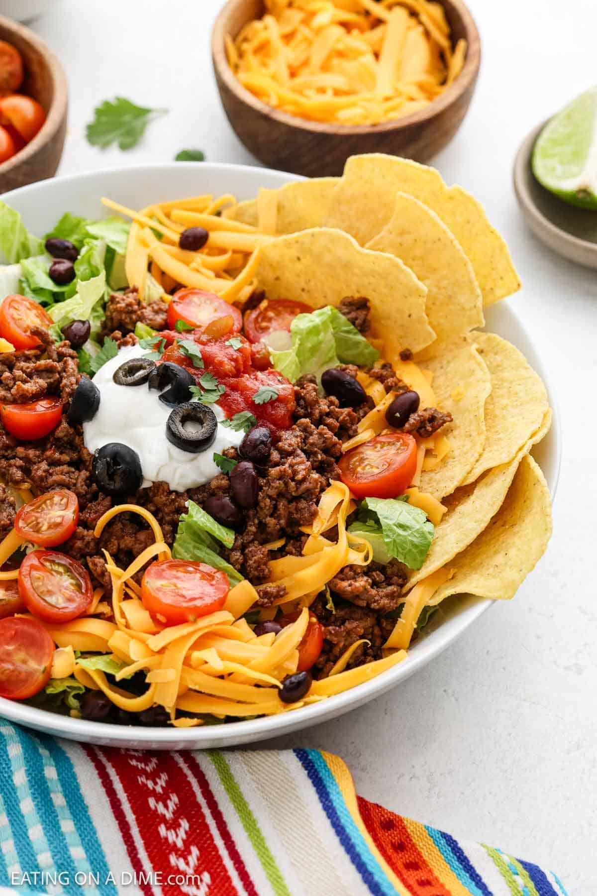 A vibrant taco salad in a white bowl featuring ground beef, shredded cheddar cheese, cherry tomatoes, black olives, black beans, lettuce, sour cream, and salsa. Tortilla chips are arranged around the edge. A colorful cloth lies beneath the bowl.