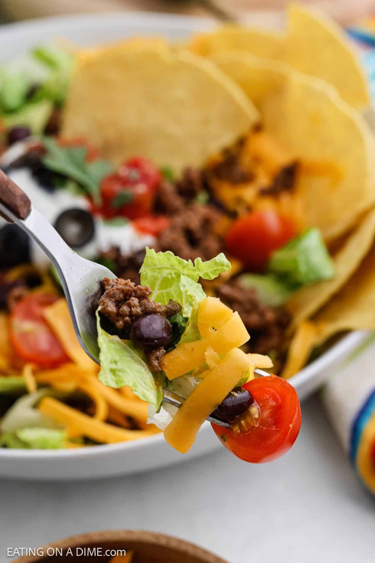 A close-up of a fork holding salad ingredients, including lettuce, ground beef, black beans, shredded cheddar cheese, and a cherry tomato. In the background, a bowl of taco salad with tortilla chips is visible.