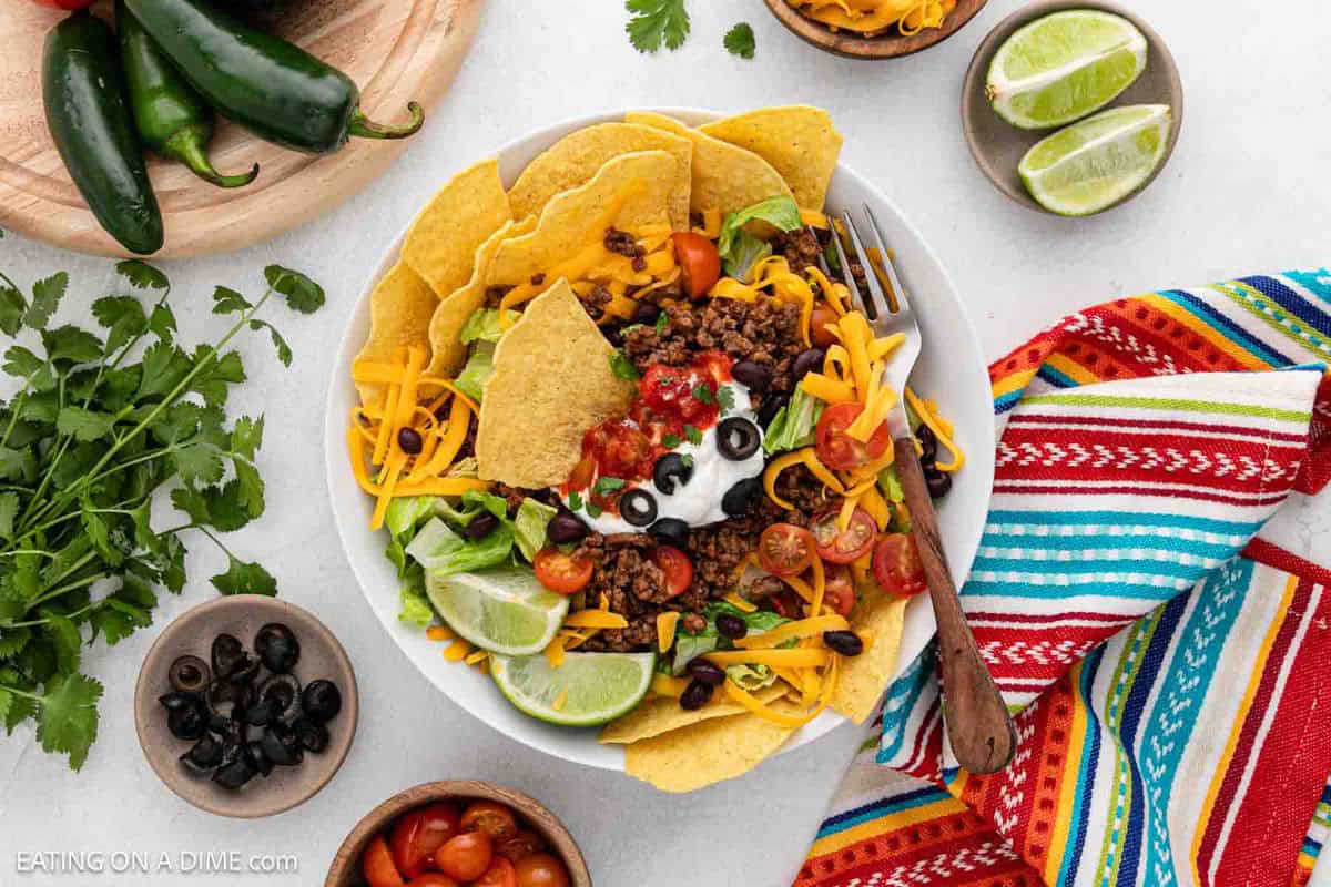 A vibrant taco salad in a white bowl, topped with tortilla chips, ground beef, cheese, sour cream, olives, tomatoes, and salsa. Lime wedges and a fork rest on the side. Surrounding are jalapeños, cilantro, black olives, and colorful striped napkins.