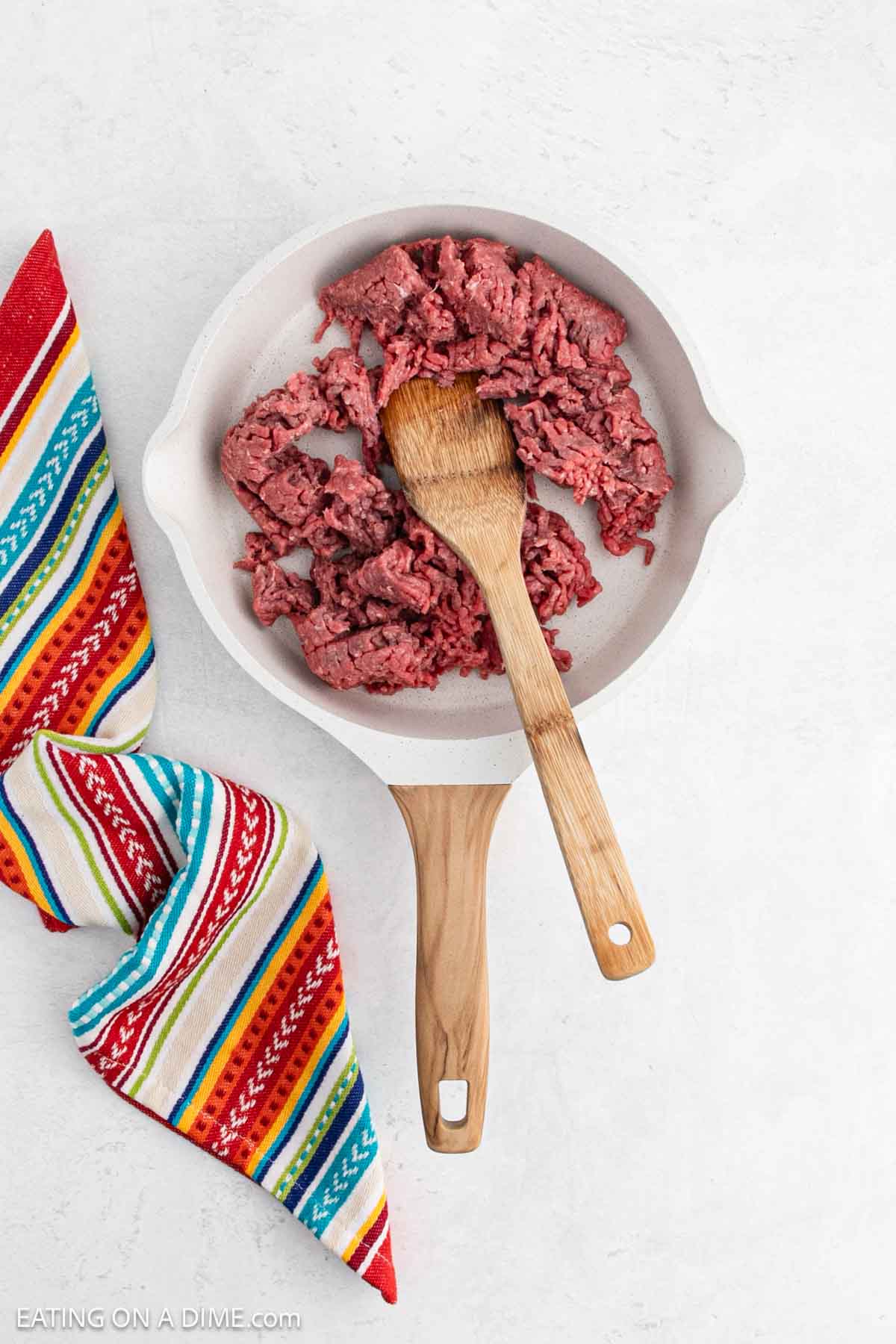 Raw ground meat in a white frying pan with a wooden handle and spoon. A colorful striped fabric is placed nearby on the light surface.