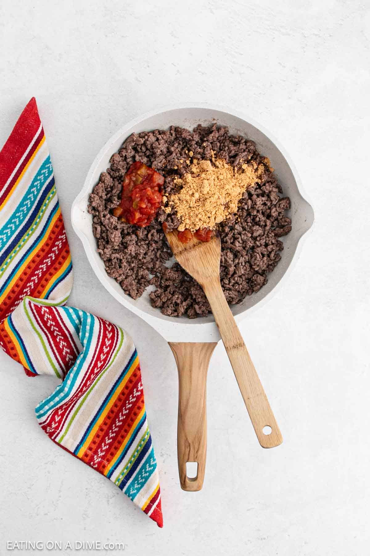A skillet with cooked ground beef topped with taco seasoning and salsa, next to a wooden spoon. A colorful, striped cloth is beside the skillet on a white surface.