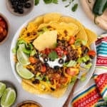 A colorful taco salad with taco chips, seasoned ground beef, shredded cheddar cheese, lettuce, cherry tomatoes, black beans, sour cream, black olives, salsa, and lime wedges, garnished with cilantro. A striped cloth and lime wedges are beside the bowl.