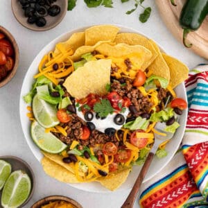A colorful taco salad with taco chips, seasoned ground beef, shredded cheddar cheese, lettuce, cherry tomatoes, black beans, sour cream, black olives, salsa, and lime wedges, garnished with cilantro. A striped cloth and lime wedges are beside the bowl.