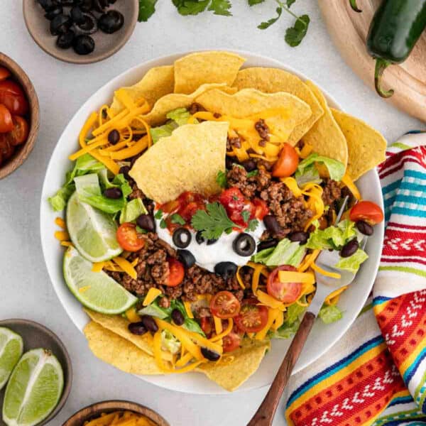A colorful taco salad with taco chips, seasoned ground beef, shredded cheddar cheese, lettuce, cherry tomatoes, black beans, sour cream, black olives, salsa, and lime wedges, garnished with cilantro. A striped cloth and lime wedges are beside the bowl.