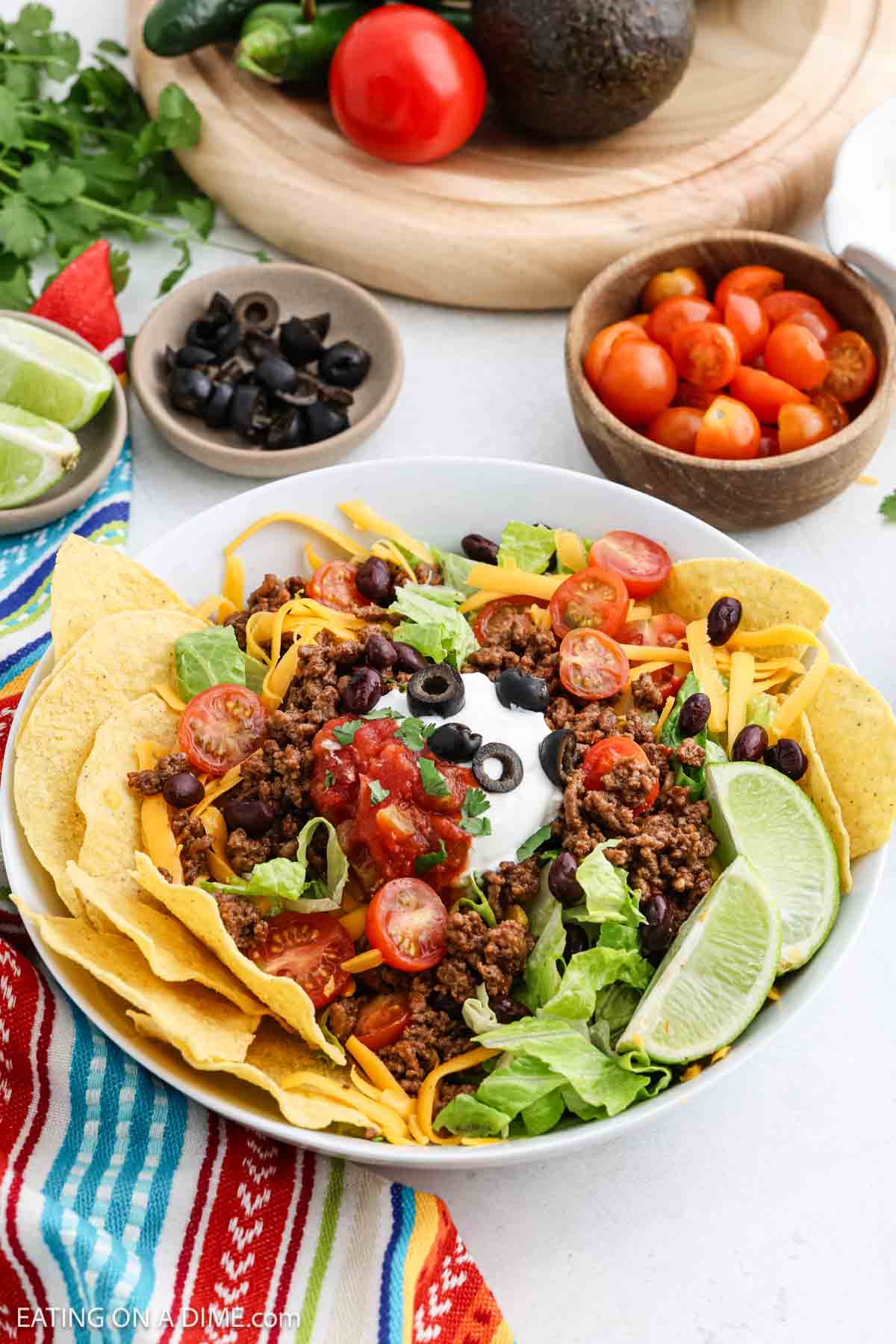 A colorful taco salad in a white bowl with ground beef, lettuce, black beans, cherry tomatoes, cheese, and sour cream, garnished with lime wedges and tortilla chips. Surrounding the bowl are small bowls of cherry tomatoes, olives, and lime wedges.