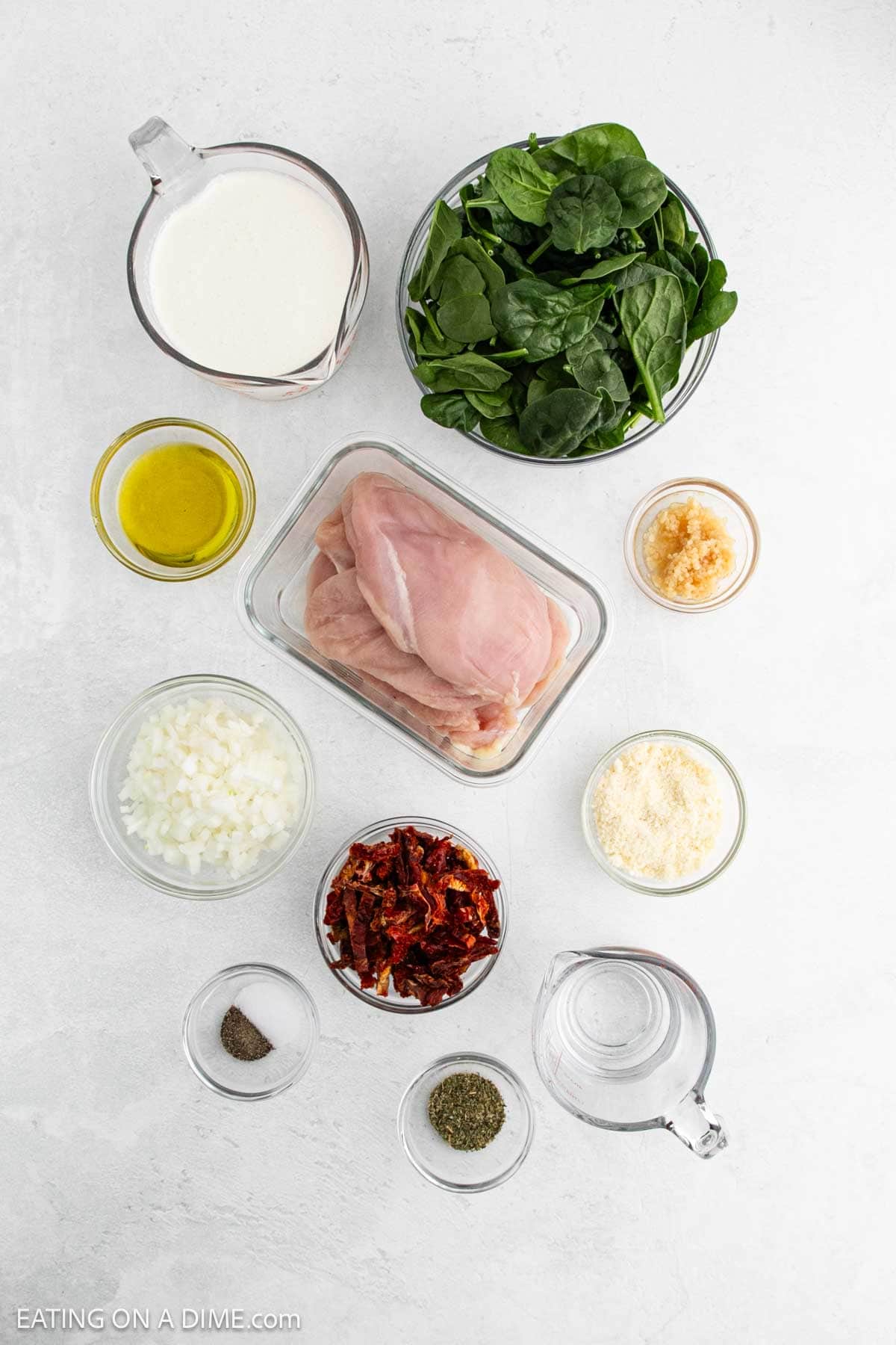 Top-down view of ingredients for a Tuscan Chicken Recipe: raw chicken breasts, fresh spinach, milk, olive oil, minced garlic, chopped onions, grated parmesan cheese, sun-dried tomatoes, black pepper, Italian seasoning, and water.