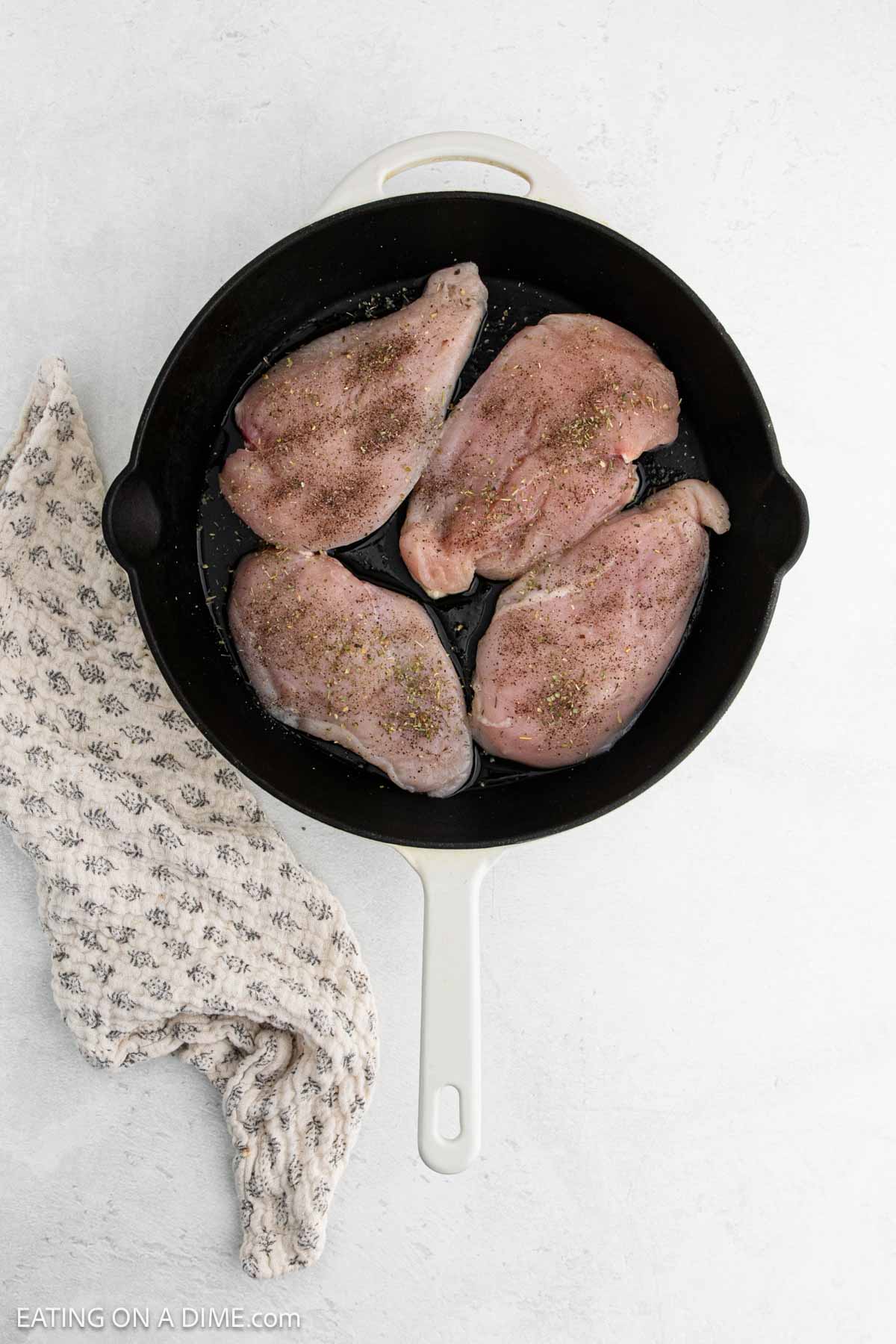 A white skillet with four raw, seasoned chicken breasts inside, ready to be transformed into a flavorful Tuscan Chicken Recipe, placed on a light surface next to a beige patterned kitchen towel.