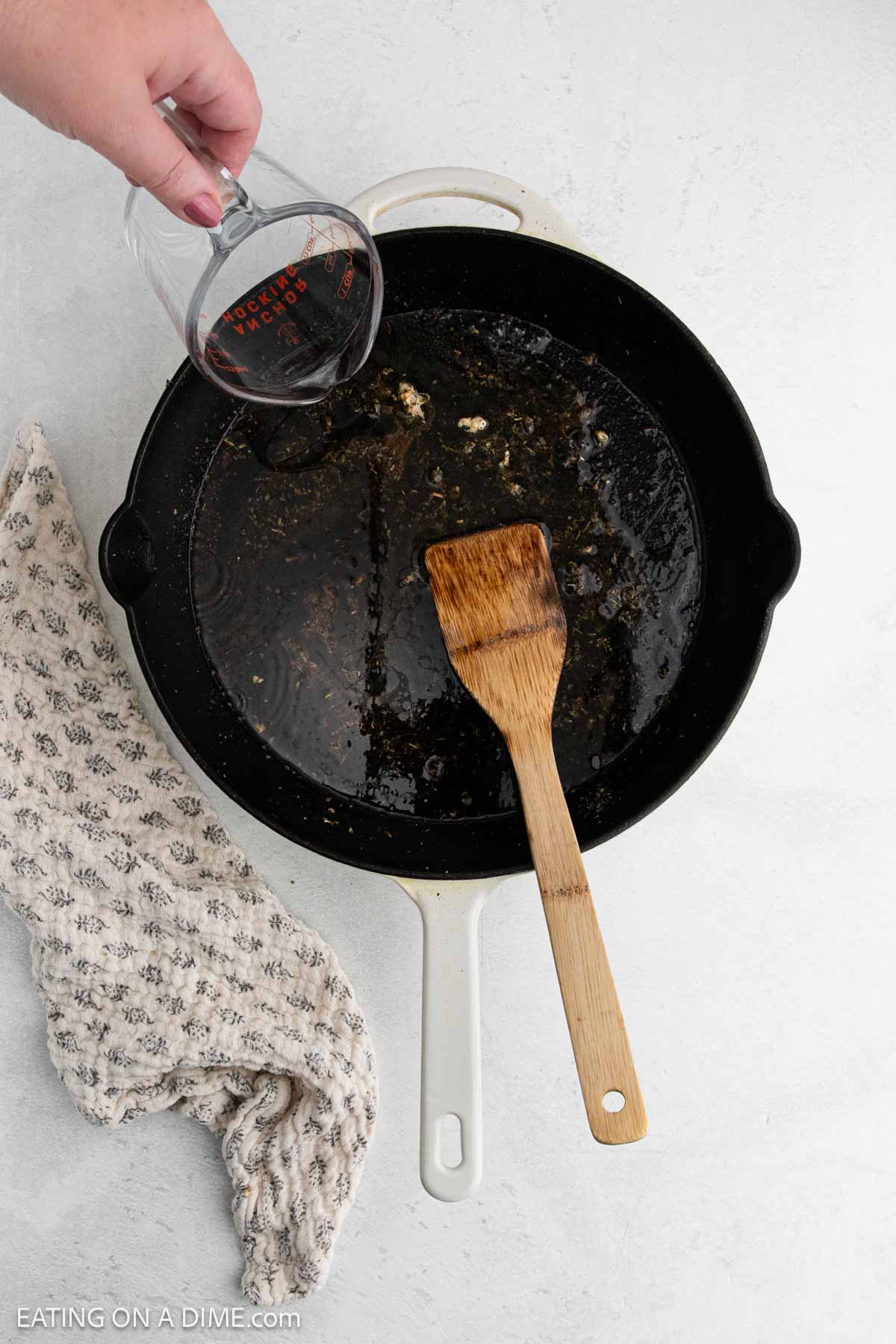 A hand pours liquid from a measuring cup into a black skillet with residue, beside a wooden spatula and a beige patterned kitchen towel—a step in preparing a delicious Tuscan Chicken Recipe.