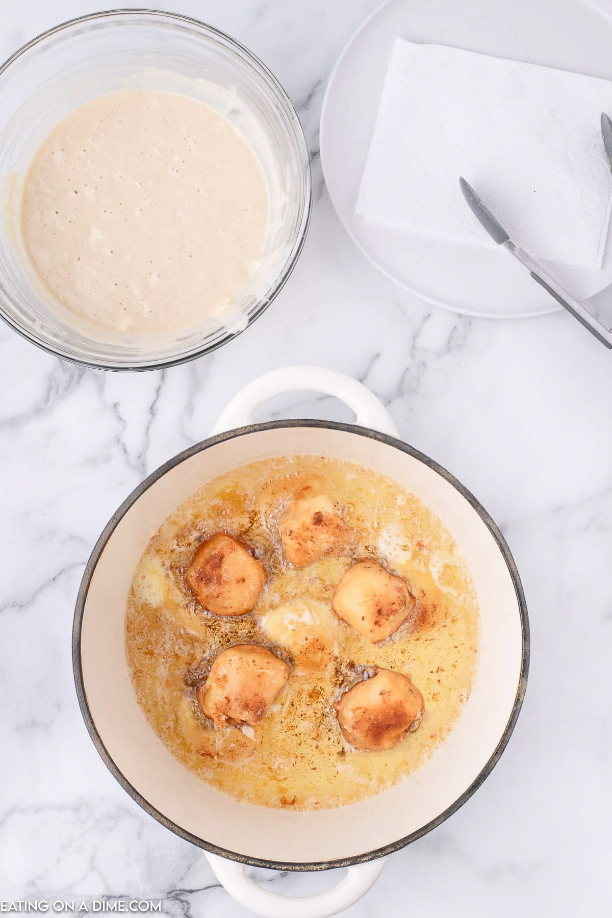 Deep fried cheesecake bites in a large pot of oil with a bowl of pancake mix on the side