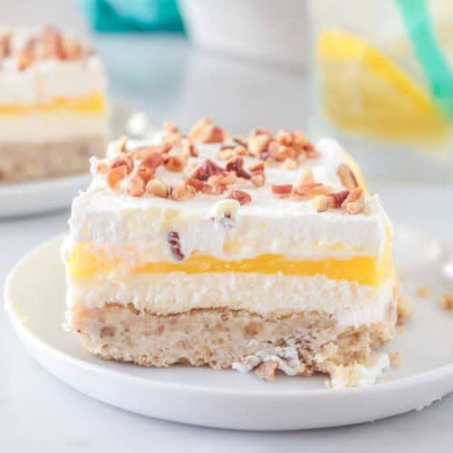A slice of Lemon Lush Dessert with a crumbly base, creamy middle, vibrant yellow lemon layer, whipped topping, and chopped nuts on top, served on a white plate.