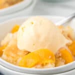 A close-up of Peach Cobbler in a bowl, topped with a scoop of vanilla ice cream, shows bright peach slices nestled within the warm dessert.