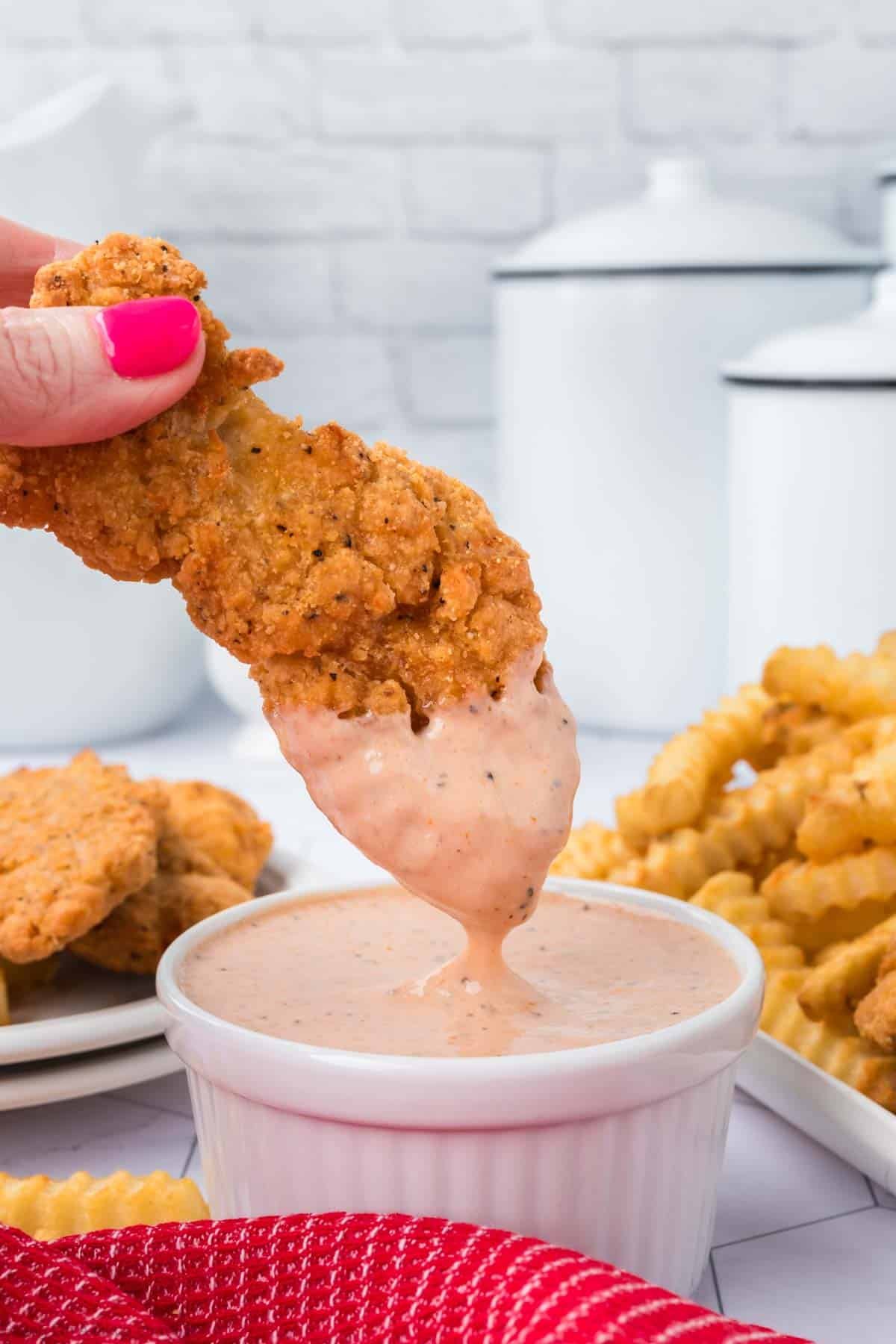 A hand with pink nail polish dips a crispy fried chicken tender into a small ramekin of creamy Raising Cane's Sauce. Crinkle-cut fries are visible in the background, set in a white kitchen setting. A red cloth rests in the foreground, capturing this delicious recipe moment perfectly.