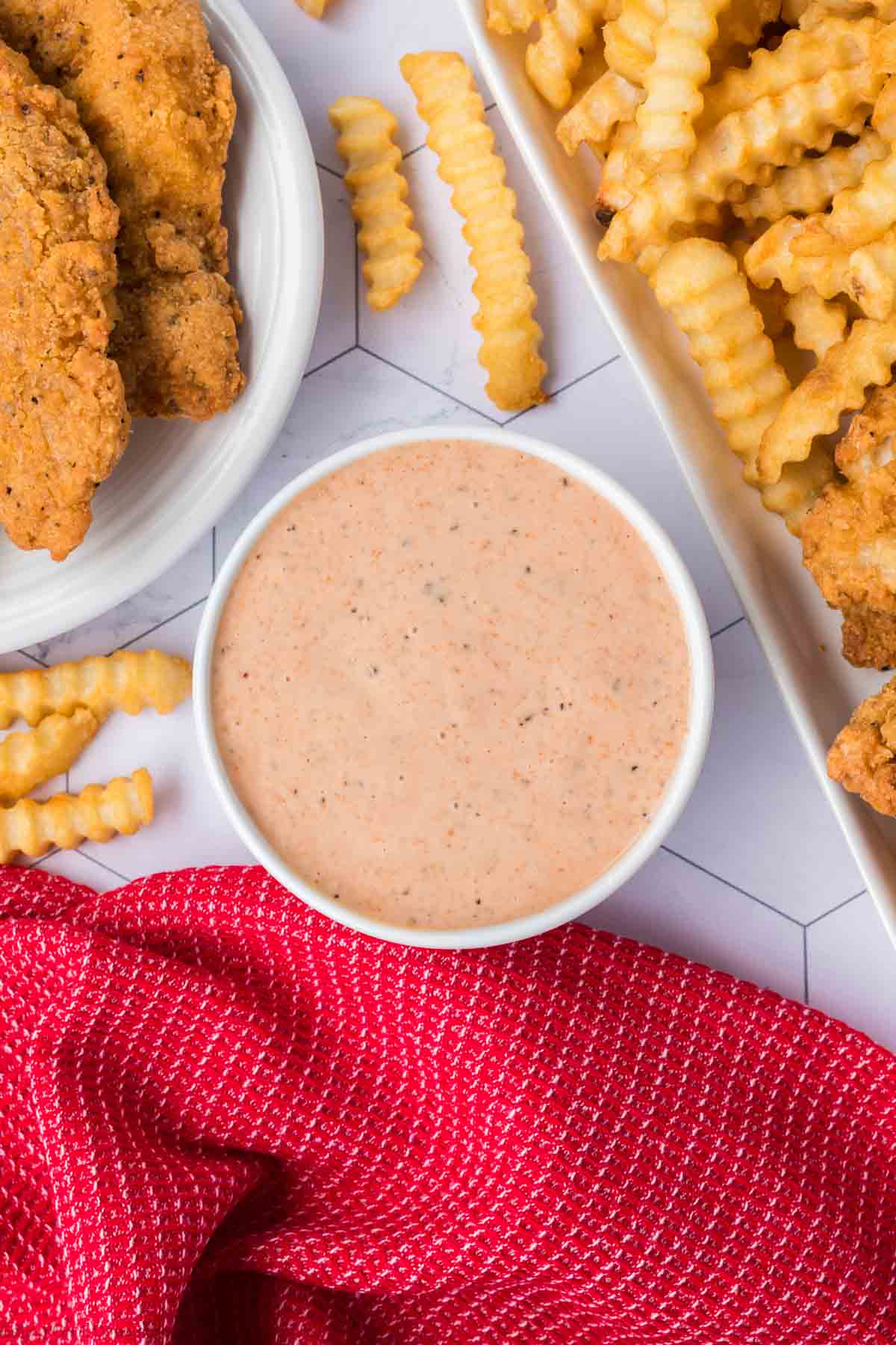 A bowl of Raising Cane's Sauce sits on a surface surrounded by crispy crinkle-cut fries and golden fried chicken. A red textured cloth is partially visible in the bottom foreground, adding a touch of warmth to this delicious scene.