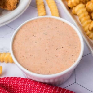 A white bowl filled with Raising Cane’s signature orange, speckled sauce rests on a white surface. Crinkle-cut fries and pieces of fried food are nearby, inviting a delicious dip. A red cloth with a textured pattern adds a splash of color in the foreground.