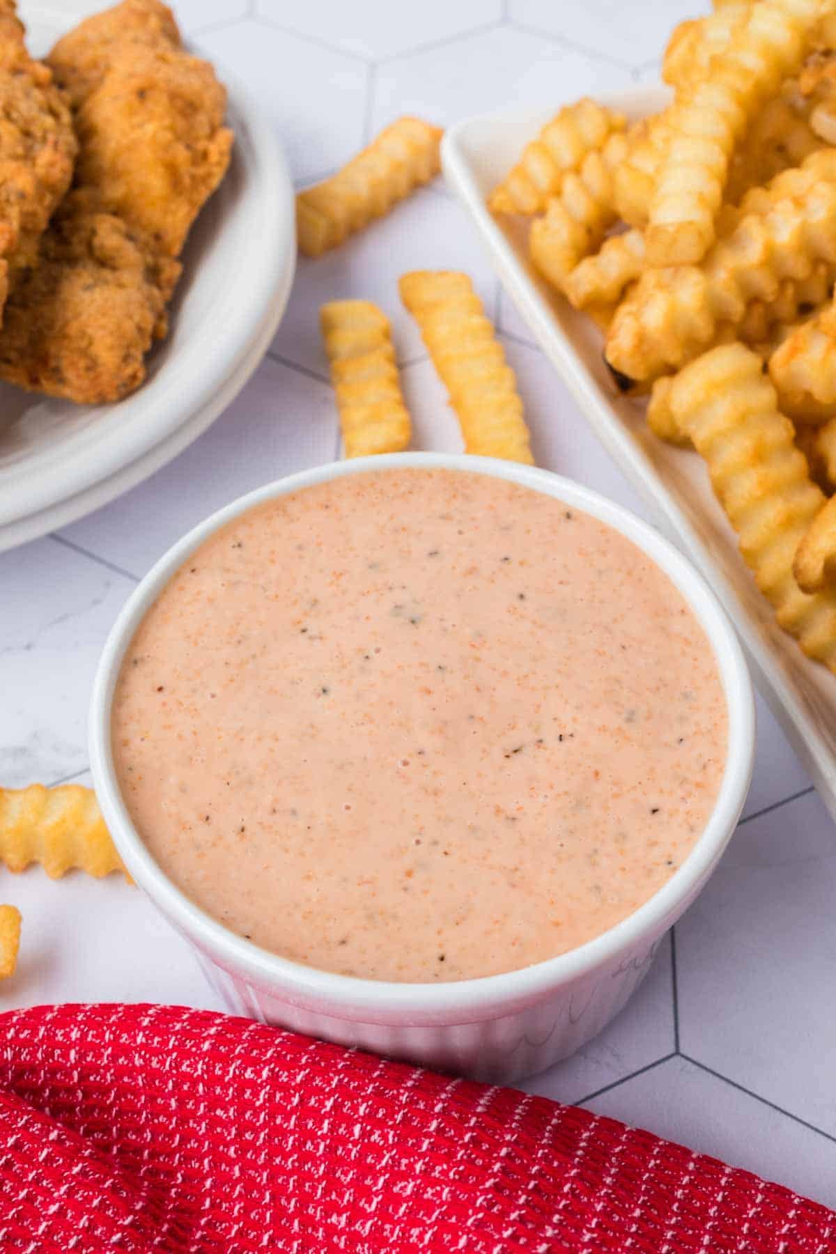 A bowl of creamy Raising Cane’s Sauce takes center stage, surrounded by crinkle-cut fries and fried chicken on a white, hexagonal-patterned table. A hint of red cloth peeks into the scene, tying together this mouthwatering recipe setup.