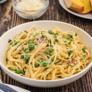 A bowl of spaghetti carbonara topped with green peas, grated cheese, and parsley. Part of Meal Plan 49, this dish sits on a rustic wooden table with a background featuring extra grated cheese and a slice of bread on a plate.