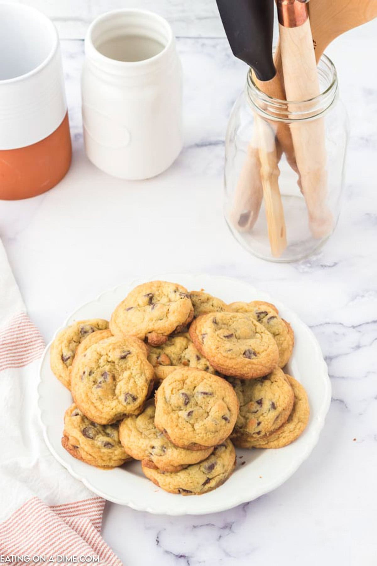 A plate of Toll House chocolate chip cookies sits on a marble countertop. Nearby, kitchen utensils in a jar and two ceramic containers add charm. A cloth napkin is partially visible on the side.