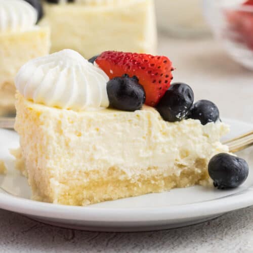 A slice of creamy cheesecake made with smooth cottage cheese is topped with whipped cream, fresh blueberries, and a halved strawberry on a white plate, with a fork nearby.