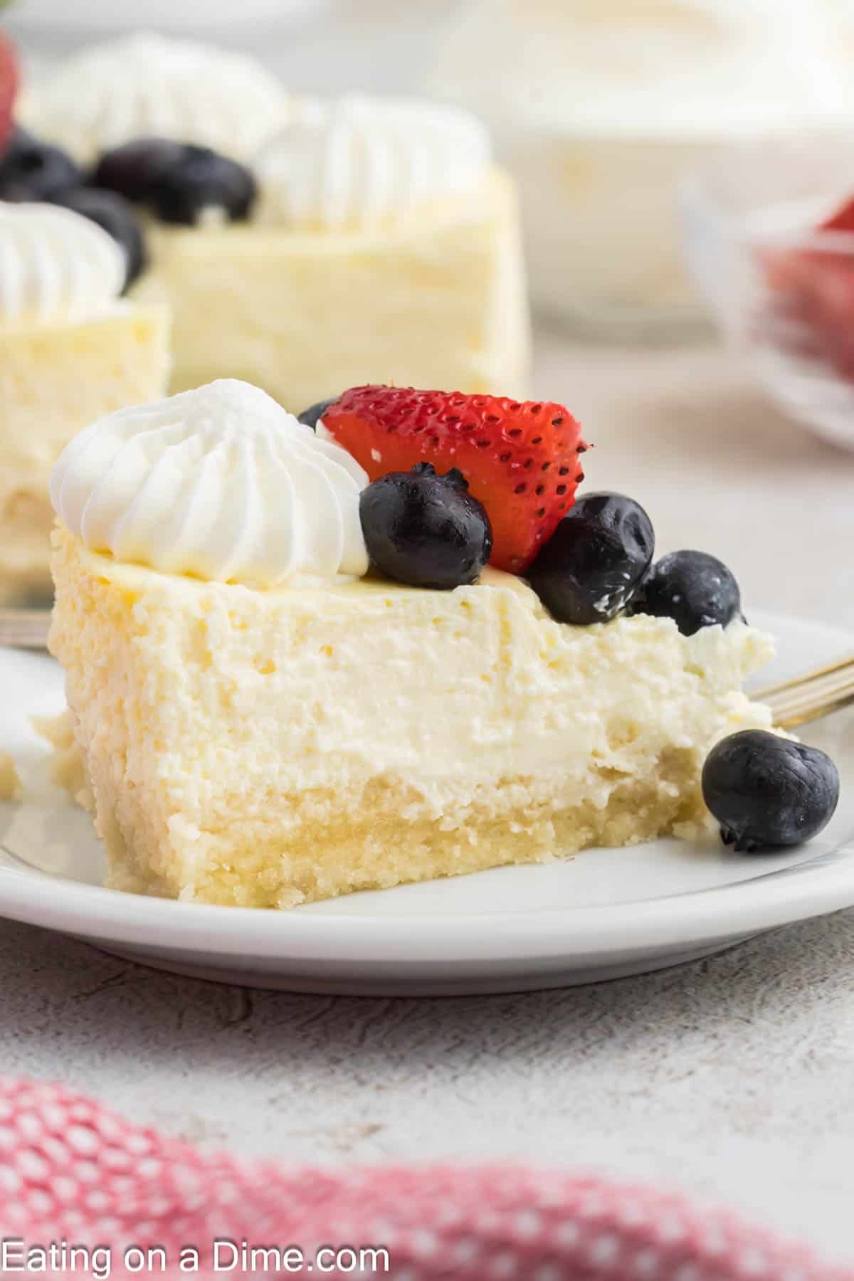 A slice of cheesecake topped with whipped cream, blueberries, and a strawberry sits on a white plate, highlighting the creamy cottage cheese base. A fork and more cheesecake are visible in the background.