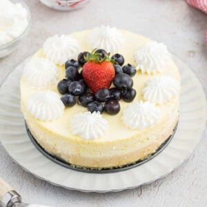 A round cottage cheese cheesecake topped with whipped cream, blueberries, and a single strawberry in the center, displayed on a white plate.