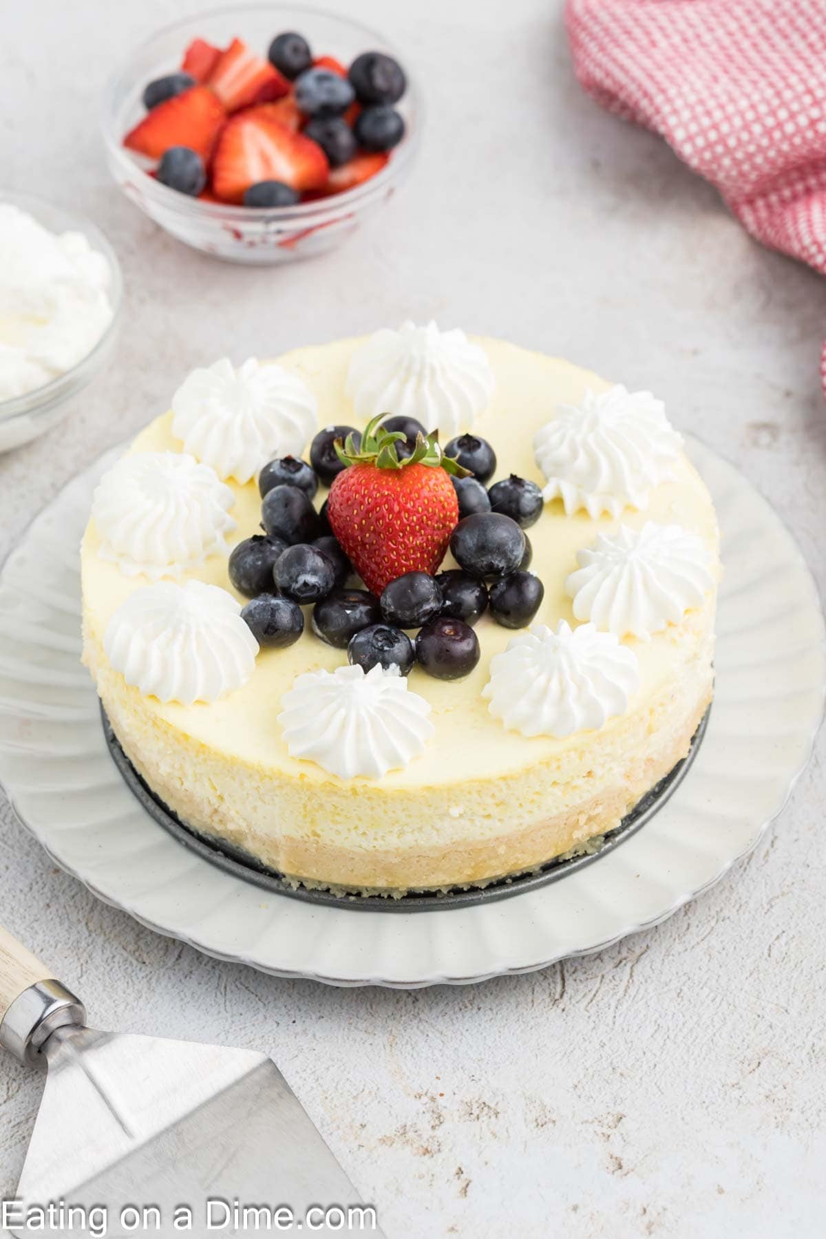 A Cottage Cheese Cheesecake topped with dollops of whipped cream, blueberries, and a large strawberry sits on a white plate. In the background are bowls of mixed berries and a red-checkered cloth.