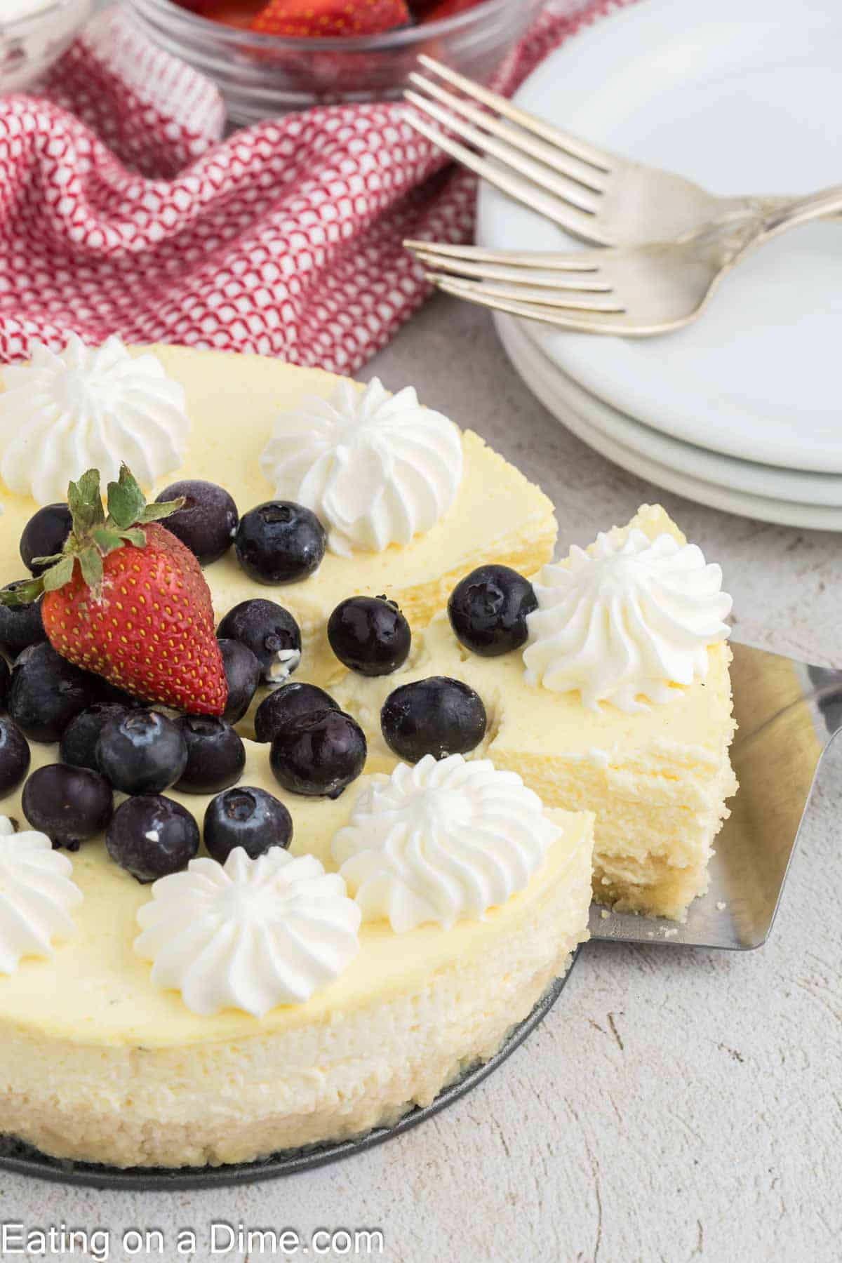 A cheesecake made with creamy cottage cheese is topped with whipped cream, fresh blueberries, and a strawberry. A slice is being lifted, while plates and forks rest in the background on a light surface.