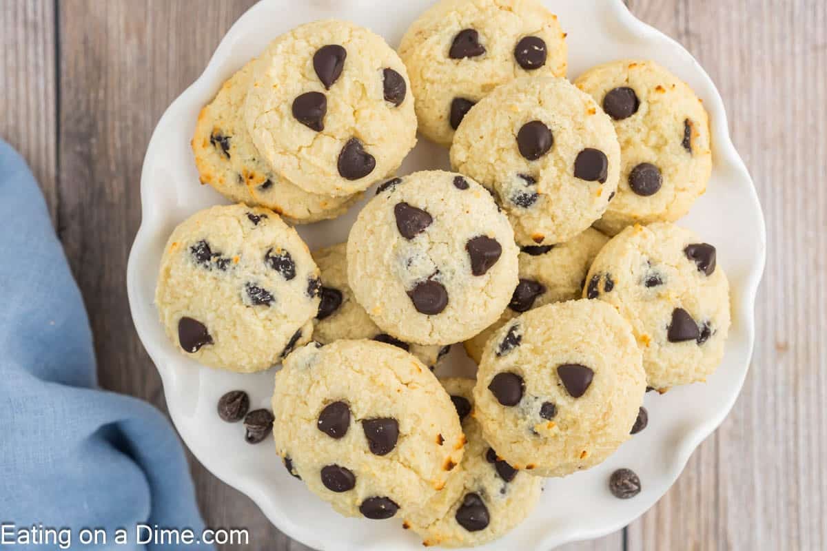 A white plate filled with homemade cottage cheese cookies sits on a wooden surface, with a few chocolate chips scattered nearby and a blue cloth partially visible to the left.