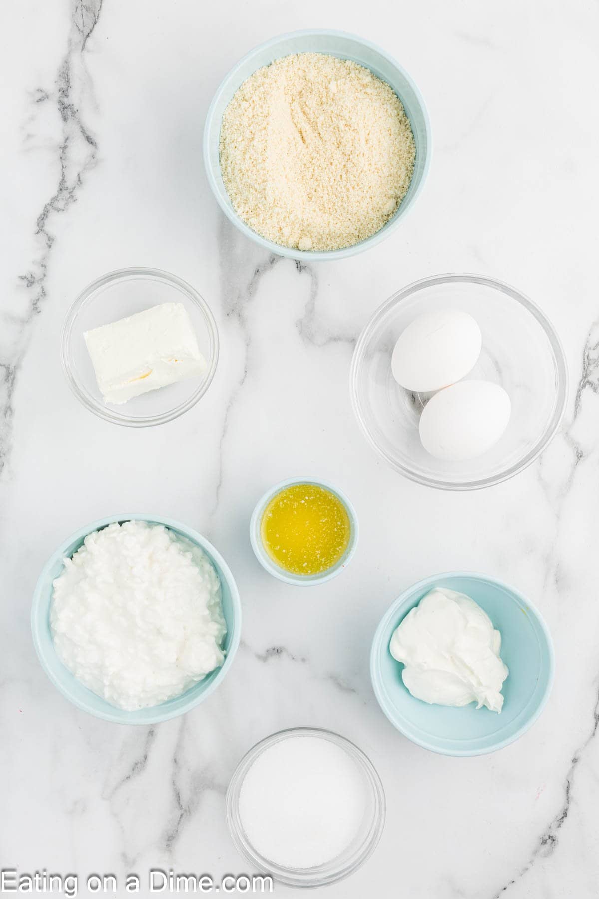 Assorted baking ingredients in small bowls on a marble surface, perfect for making cottage cheese cheesecake, including eggs, almond flour, cottage cheese, cream cheese, melted butter, sour cream, and sugar.