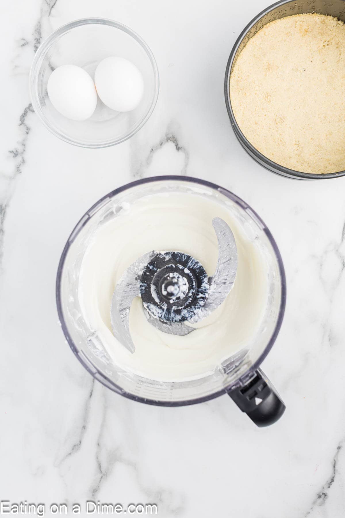 A food processor with creamy cheesecake mixture inside sits on a marble countertop, next to a bowl with two eggs and a round pan of graham cracker crumbs—perfect for making cottage cheese cheesecake.