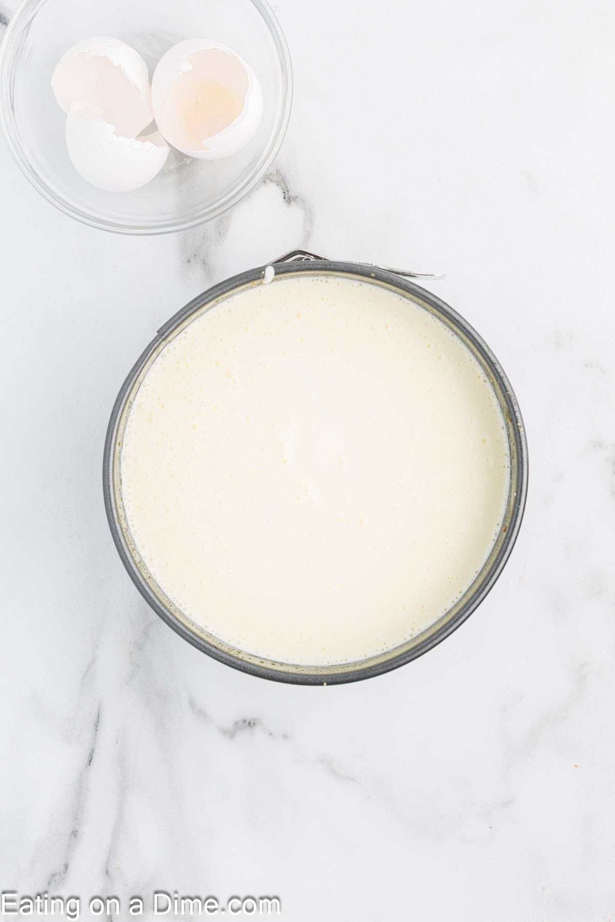 A round pan filled with smooth, pale Cottage Cheese Cheesecake batter sits on a marble surface next to a small glass bowl containing cracked eggshells. "Eating on a Dime.com" is visible at the bottom of the image.