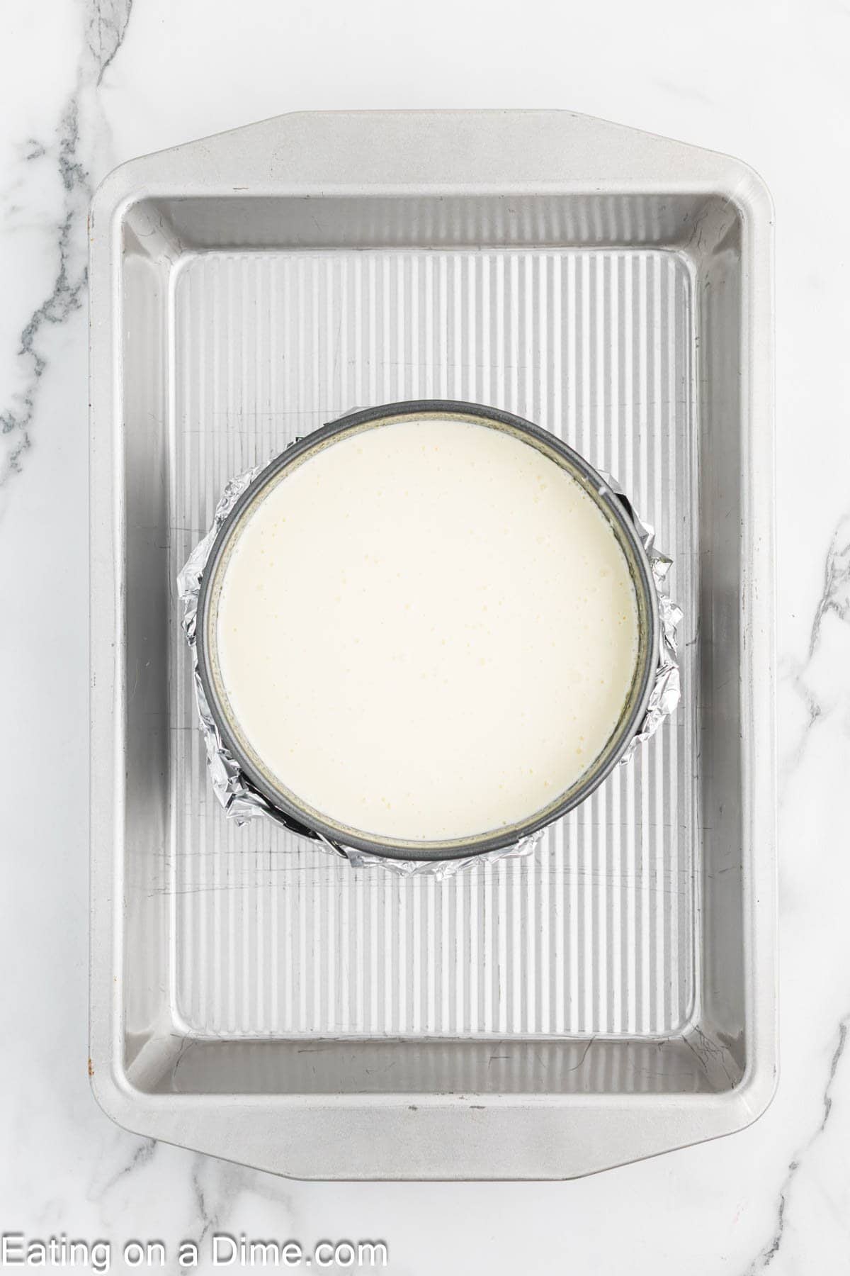 A round cake pan wrapped in foil is filled with creamy cheesecake batter and placed inside a larger rectangular baking pan on a marble surface. The bottom left corner reads “Eating on a Dime.com.”.