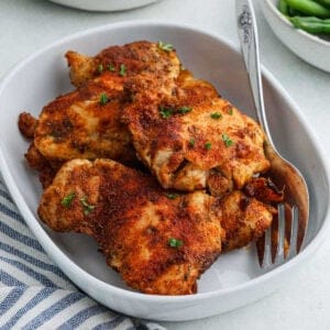 A white dish contains several pieces of seasoned, golden-brown air fryer chicken thighs, garnished with chopped herbs. A fork rests on the side of the dish, and a striped cloth is nearby—perfect for fans of easy air fryer recipes.